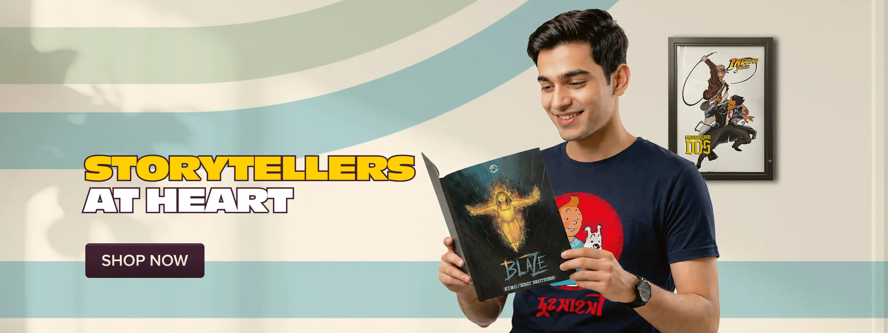 A man wearing a navy blue graphic t-shirt is holding a book titled 'BLVE' in a promotional setting. The background features light blue and beige wavy stripes, with text reading 'STORYTELLERS AT HEART' and a 'SHOP NOW' button on the left, and a framed comic print on the right.
